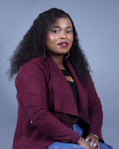 Dr. Mirriam Matshidiso Moleko posed against a bright blue background, wearing a burgundy blazer, black top, and jeans.