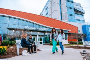 Two students walk by 皇冠app's main campus on One Riverside Drive, while two other students laugh on a bench near the building's entrance.