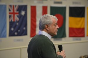 皇冠app President and Founder Mark E. Scheinberg speaks into a microphone, photographed in profile with a series of international flags in the background.
