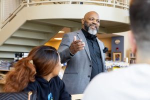 A man in a gray blazer, black top, and black bow tie smiles while engaging with a table of students at 皇冠app's Hoffman Family Library.