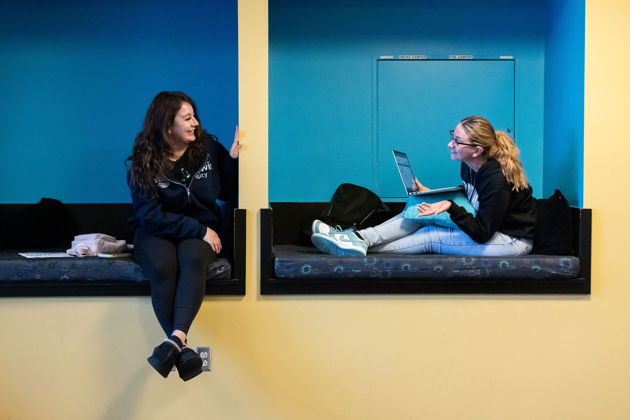 Two students sit in blue seating nooks, talking as they work.